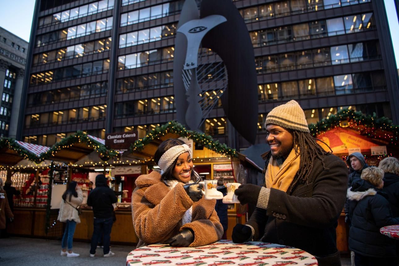 Two people chat at an outdoor holiday market stall in front of a lit building at dusk.