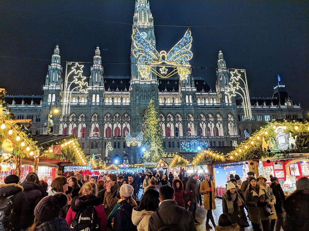 Night market crowd before an illuminated historic city hall with a tall lit Christmas tree.