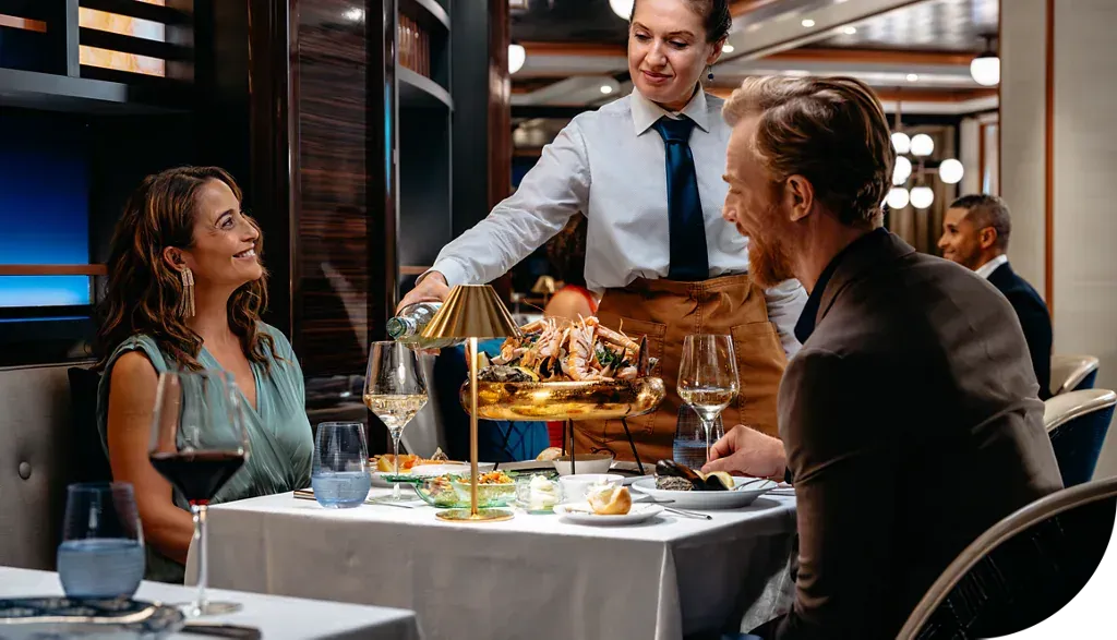 A server pours wine for a couple dining at a table in a softly lit, upscale restaurant.