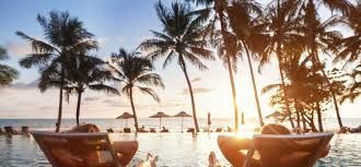 Two people relaxing in lounge chairs at an infinity pool overlooking a tropical beach at sunset.