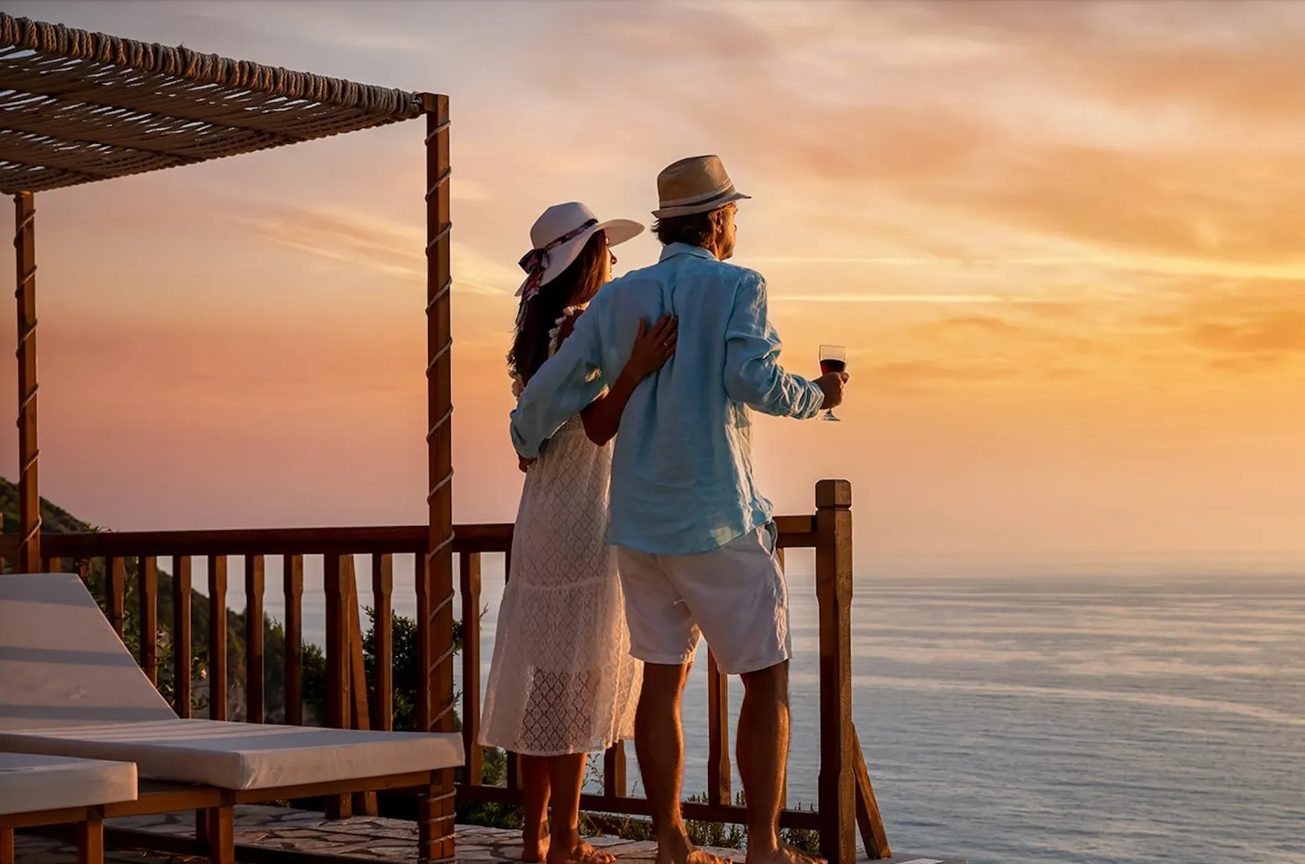 A couple in hats stands on a balcony overlooking the ocean at sunset, holding glasses of wine.