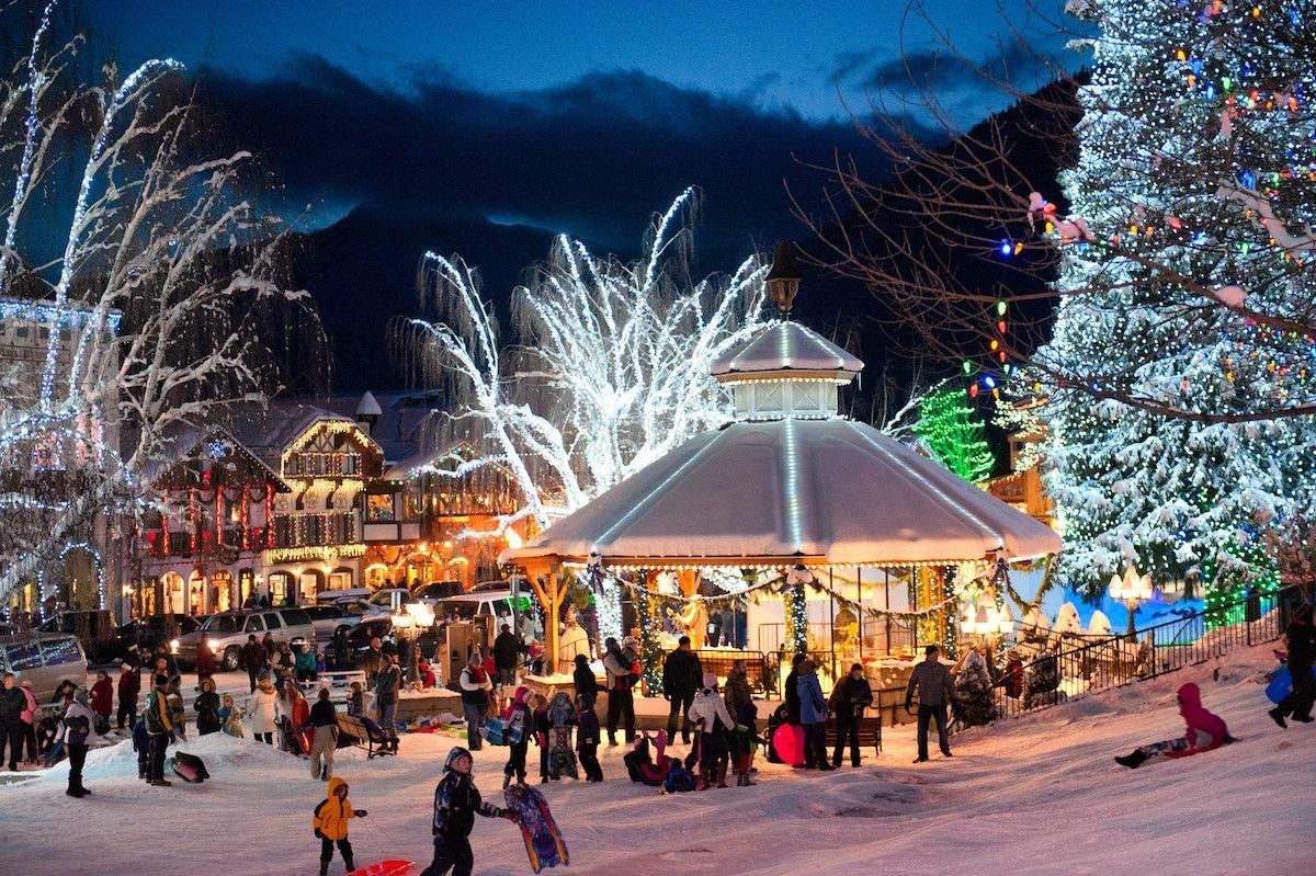 Crowded snowy village square with a lit gazebo, decorated trees, and people ice skating at dusk