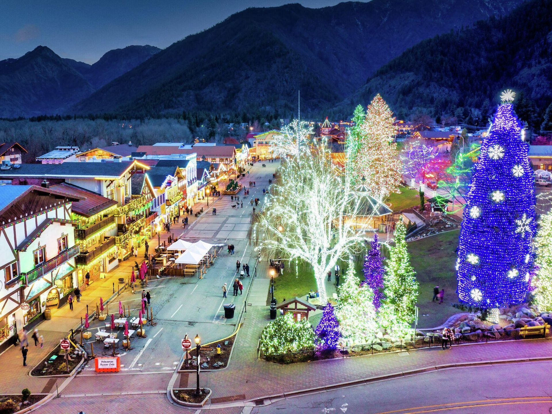 Festive plaza at dusk with colorful holiday lights, decorated trees, and mountain backdrop