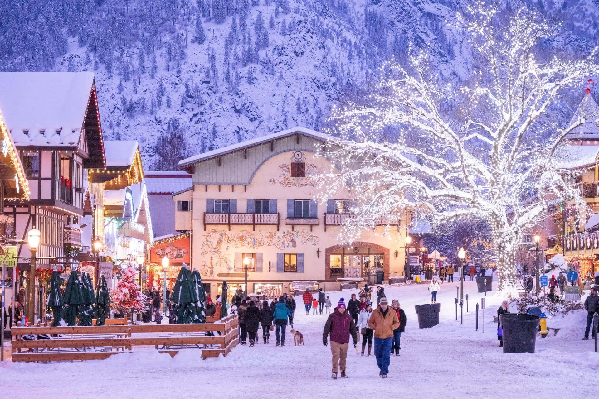Snowy village square with people walking, lit shops, and a glowing tree at dusk