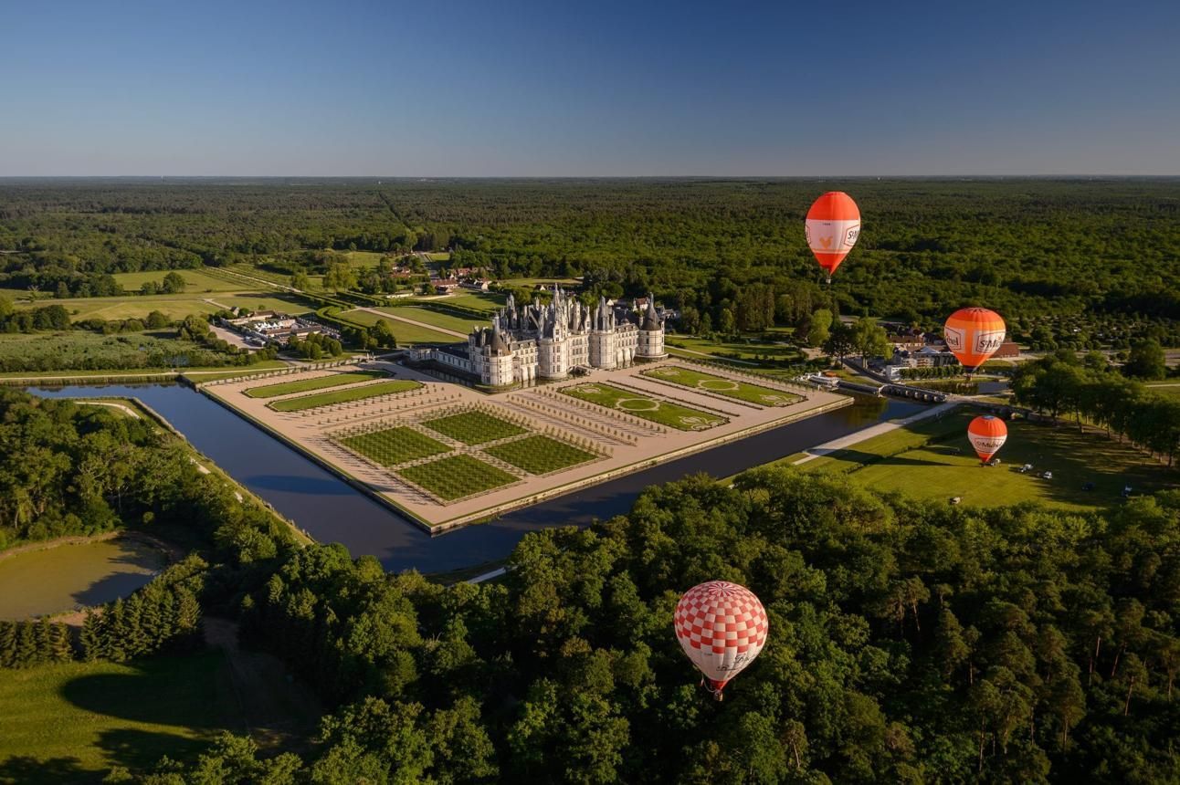 Aerial view of a castle-like estate in forested fields with several hot air balloons floating nearby