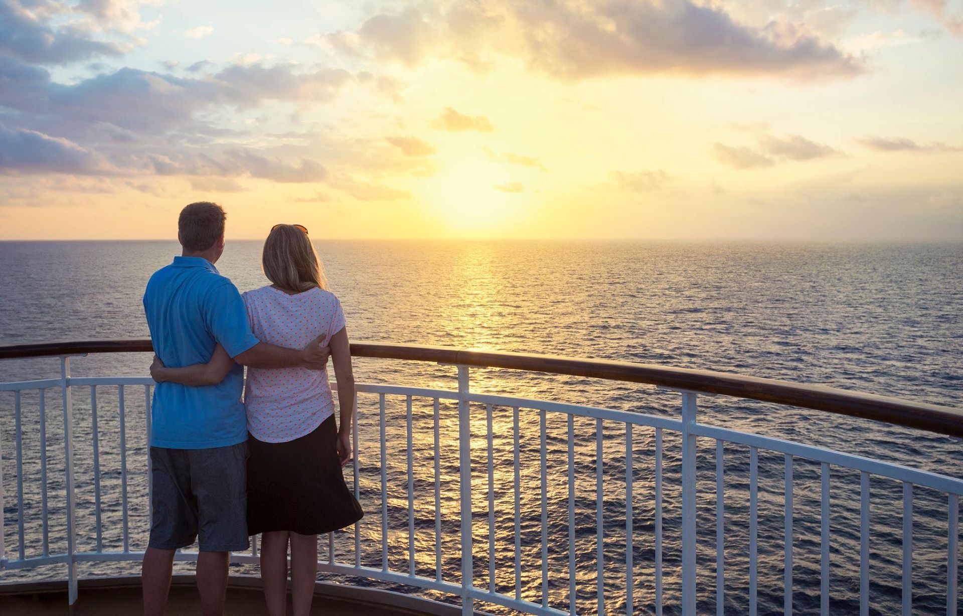 A couple stands arm-in-arm on a cruise ship deck, looking out at the sun setting over the ocean.