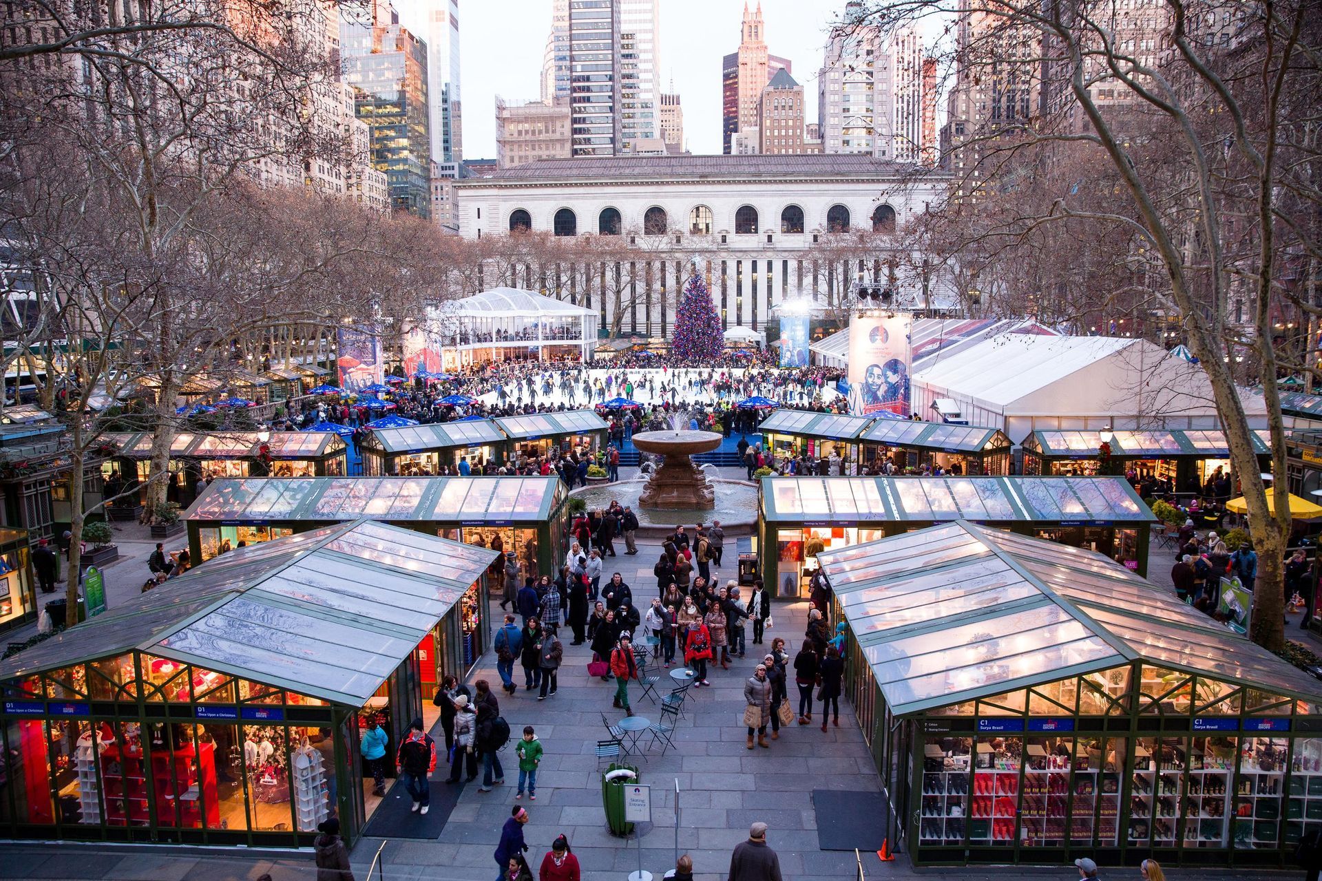 Crowded outdoor holiday market with lit stalls and a city skyline in the background