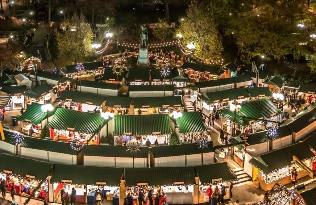 Nighttime outdoor market with green-and-white tents, bright lights, and crowds in a circular plaza.