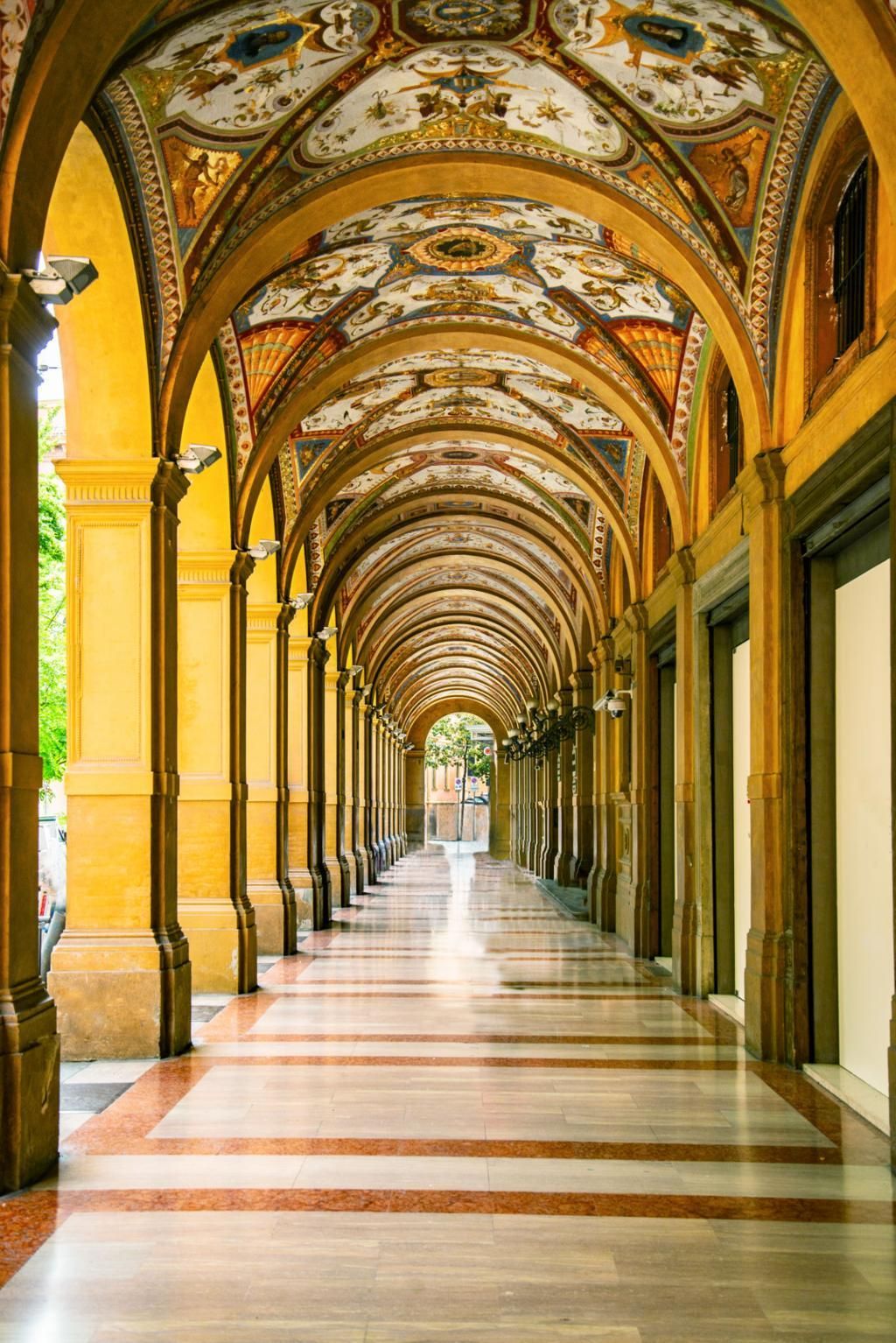 Ornate arched colonnade with yellow columns, painted vaulted ceiling, and a long tiled corridor.