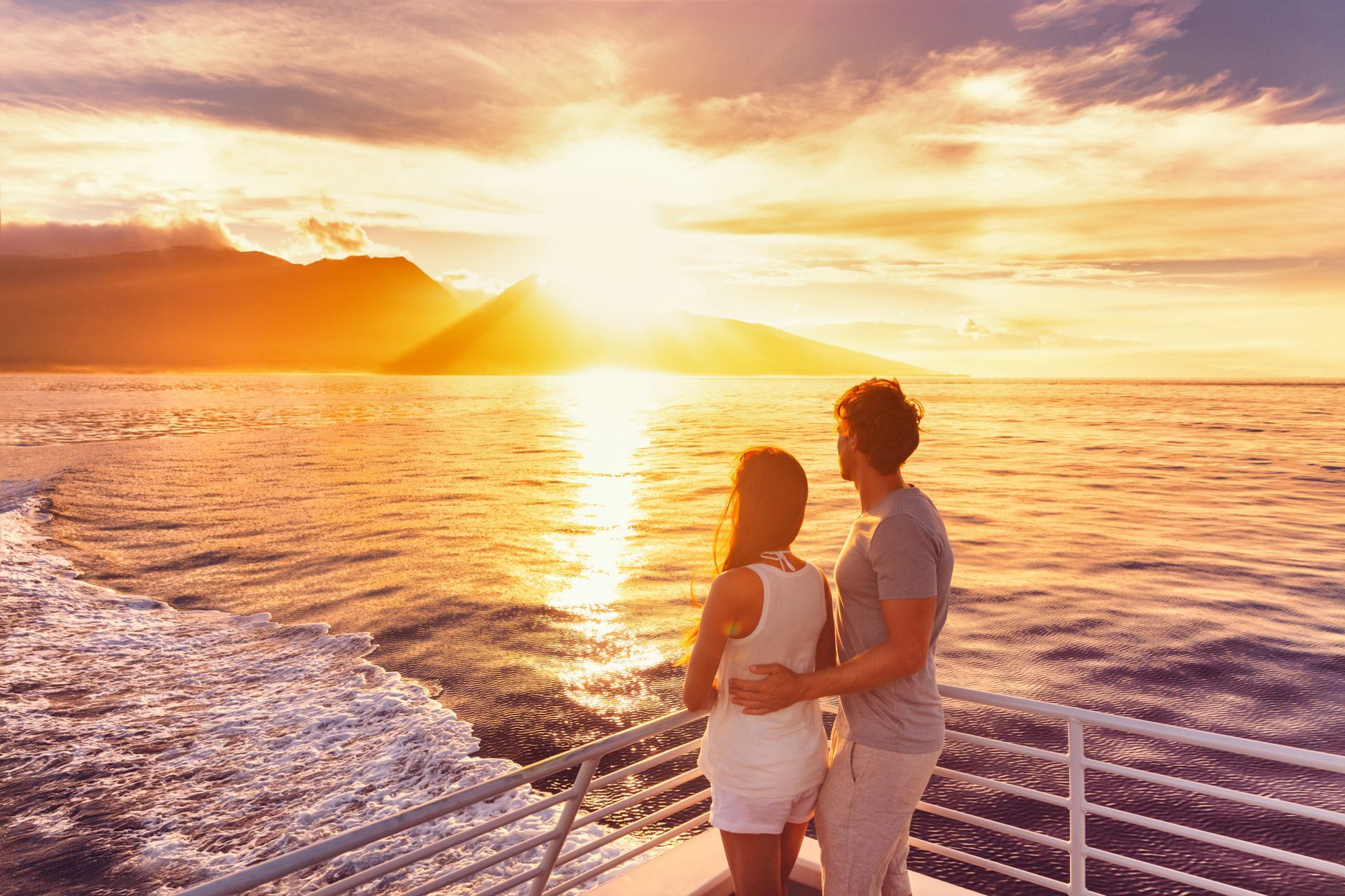 A couple stands on a boat deck, gazing at a vibrant sunset over the ocean and mountains.