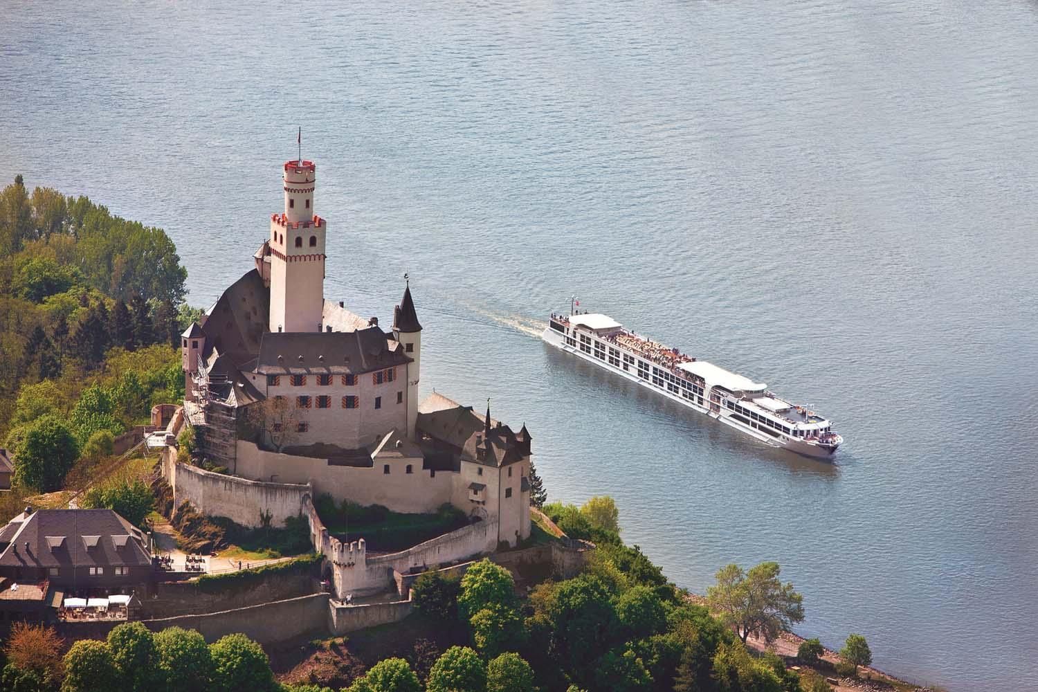 A white castle on a lakeside cliff with a tour boat passing on calm blue water.