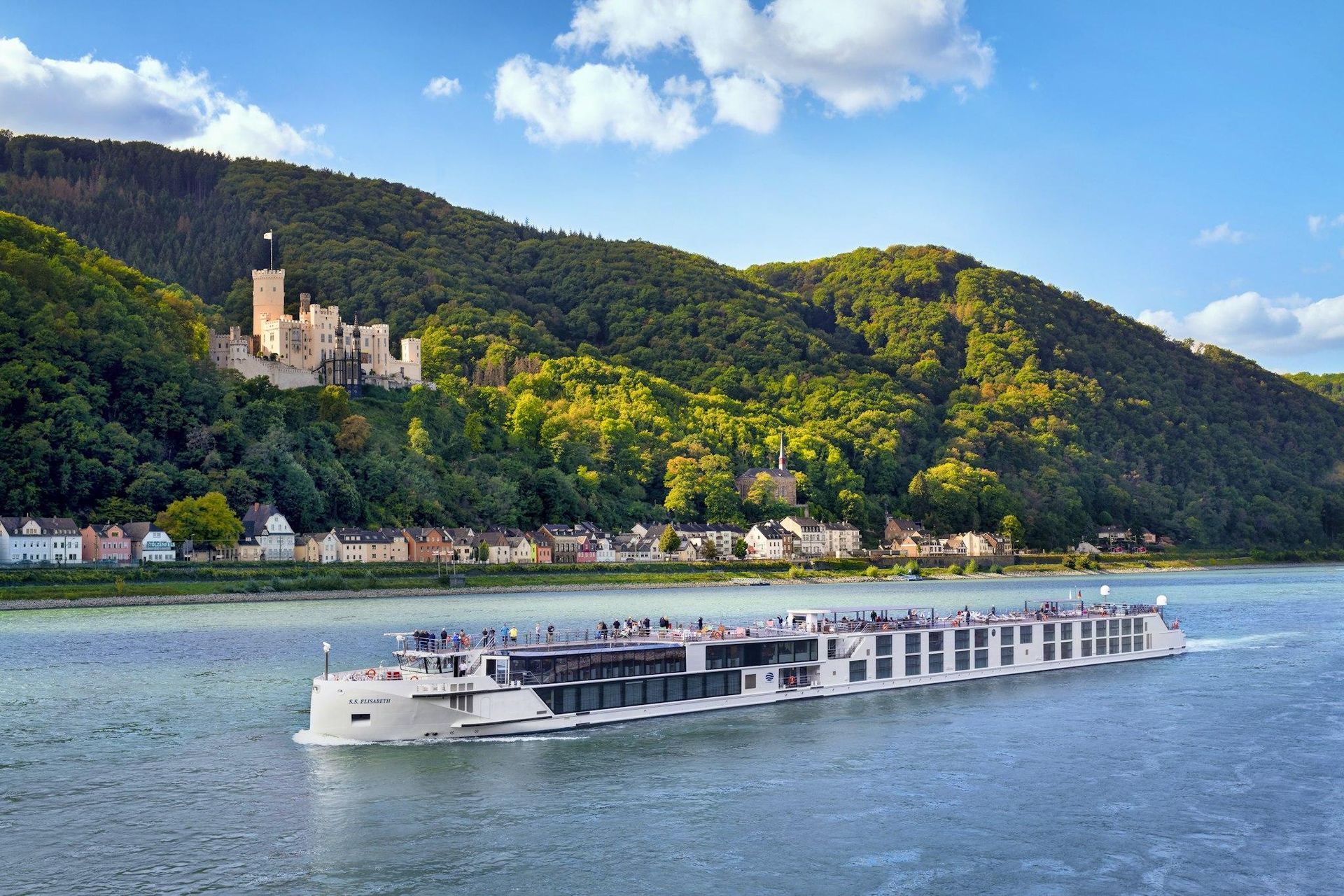 River cruise boat on a wide blue river with a forested hillside, castle, and village along the shore