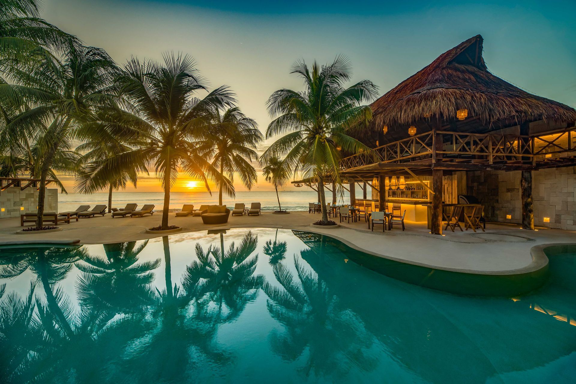 A sunset glows over a tropical resort, with palm trees and an open-air pavilion reflected in a still swimming pool.
