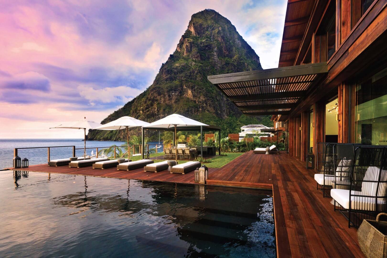 An infinity pool on a wooden deck overlooks the ocean and a large, iconic volcanic peak under a sunset sky.