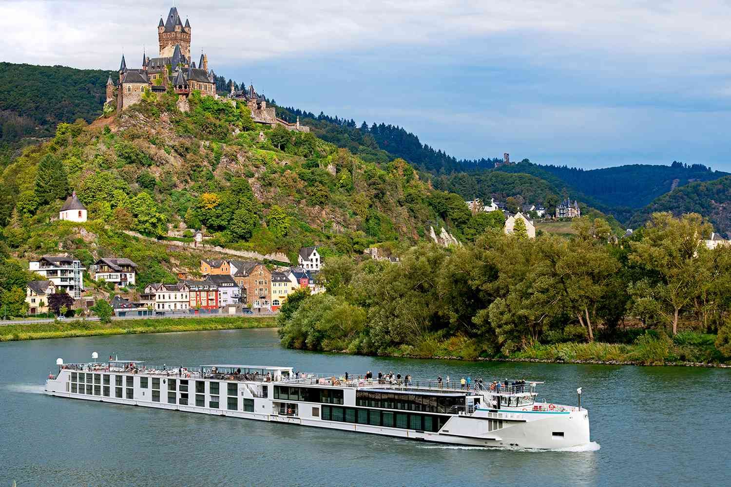 A white river cruise ship travels along a scenic river, passing a small town and the historic Cochem Castle on a hill.
