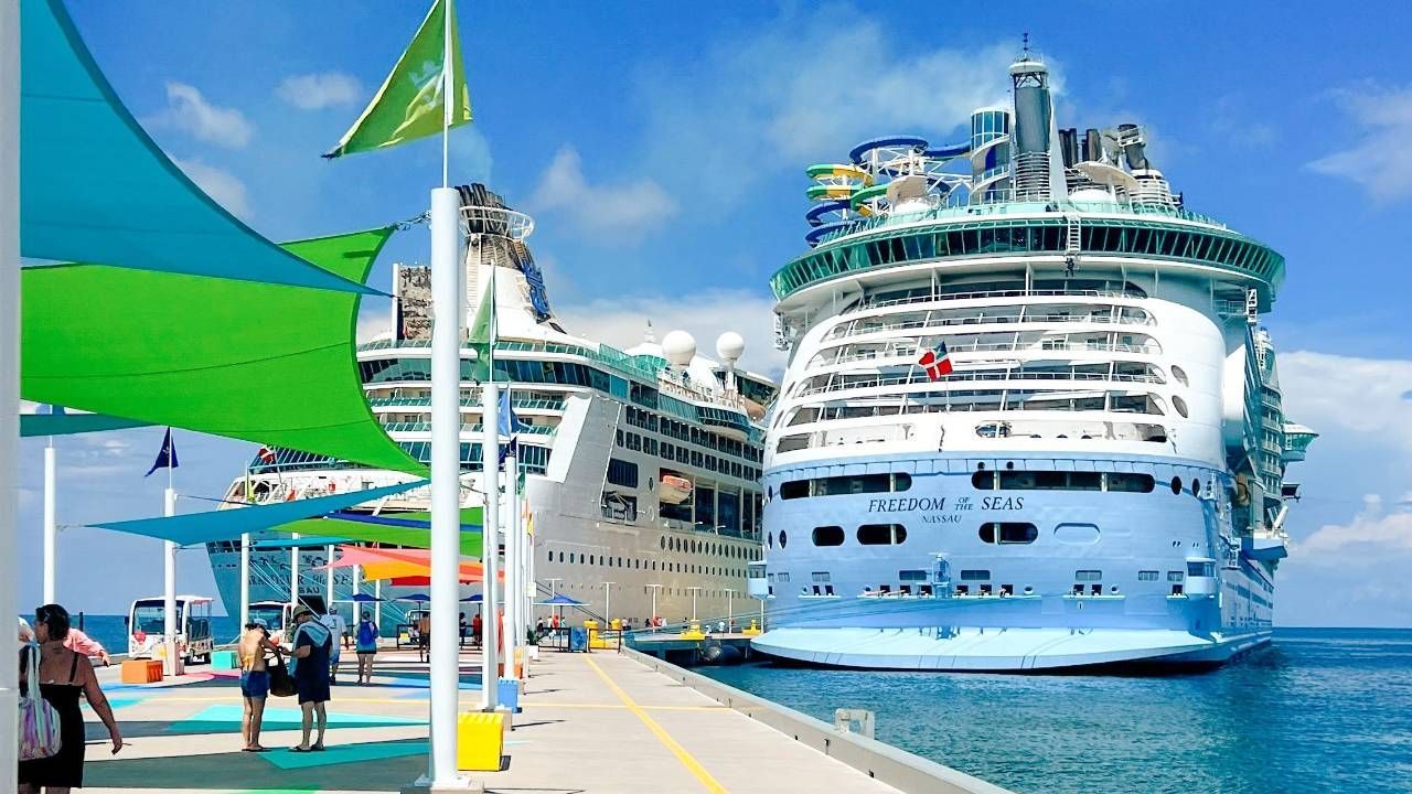 Two large Royal Caribbean cruise ships docked at a pier under a sunny sky with green and blue overhead sunshades.