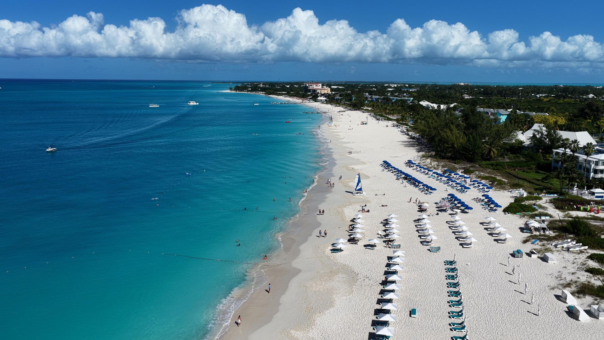 Aerial view of a long, white sand beach with rows of umbrellas and lounge chairs along a vibrant turquoise ocean.