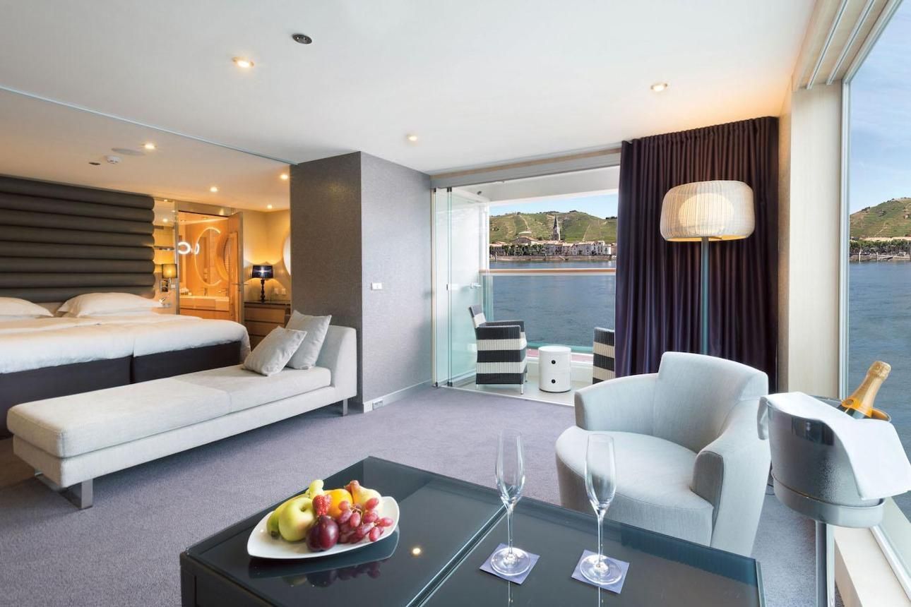 Modern hotel room with king bed, sofa, dining area, and floor-to-ceiling windows overlooking water.