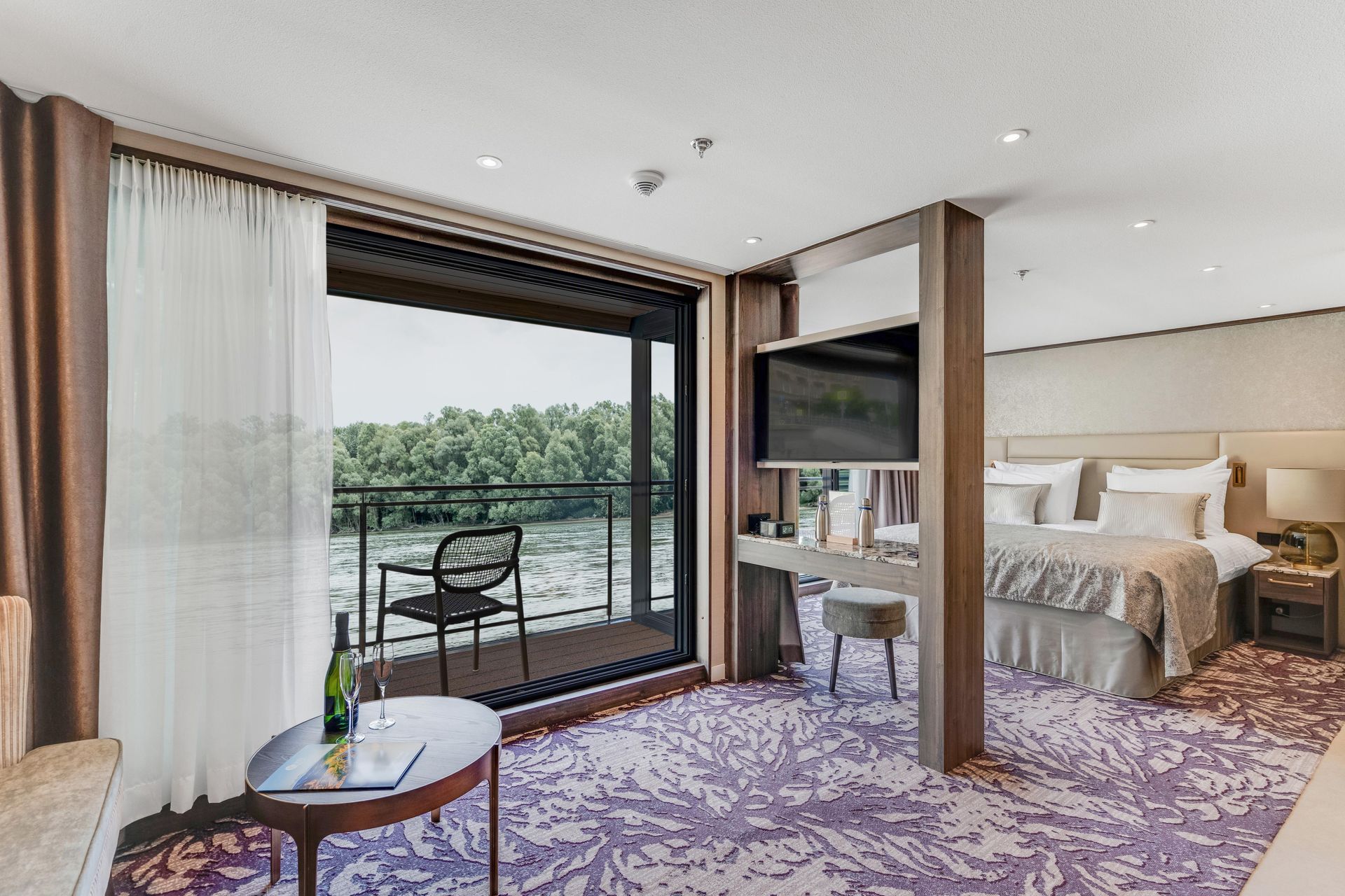 Modern hotel room with king bed, TV, lounge chair, and balcony overlooking water and trees