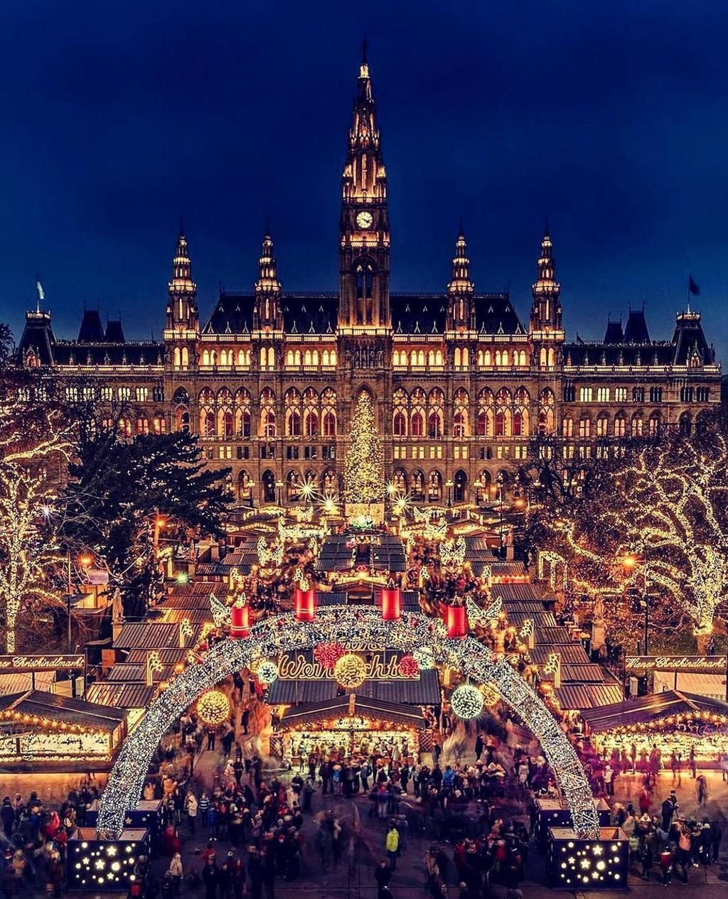 Nighttime holiday market in front of a lit historic building, with a decorated central tree and crowds below