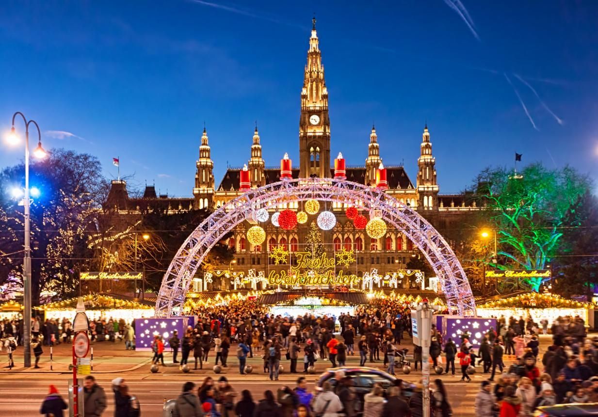 Crowd at a festive illuminated plaza with an arch of lights and a lit clock tower at dusk