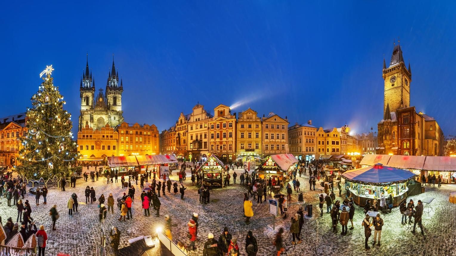 Crowded Christmas market at dusk with a lit tree, stalls, and historic buildings in a city square