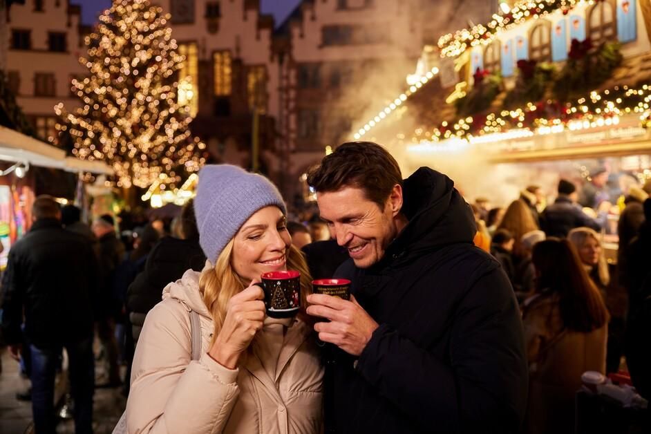 Couple smiling and clinking mugs at a festive outdoor holiday market at night