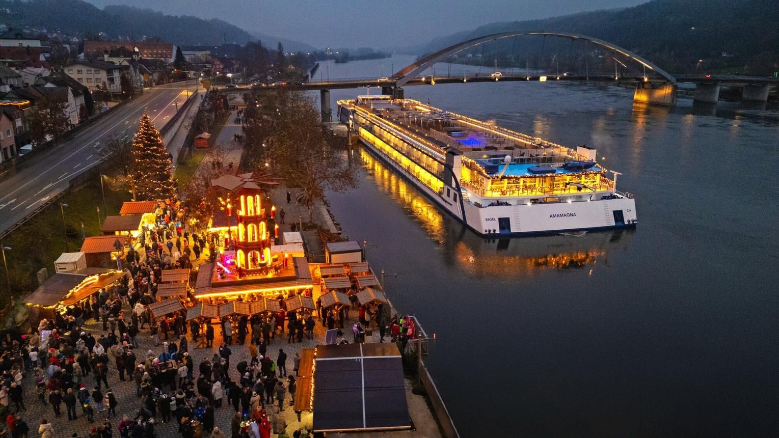 Riverfront festival at dusk with a lit boat cruise passing a crowded dock and a bridge in the background