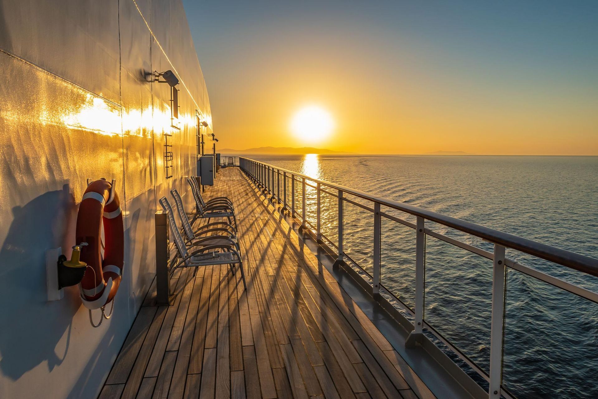 A sunrise viewed from the wooden deck of a ship, with a life ring on the left and deck chairs stretching into the distance.