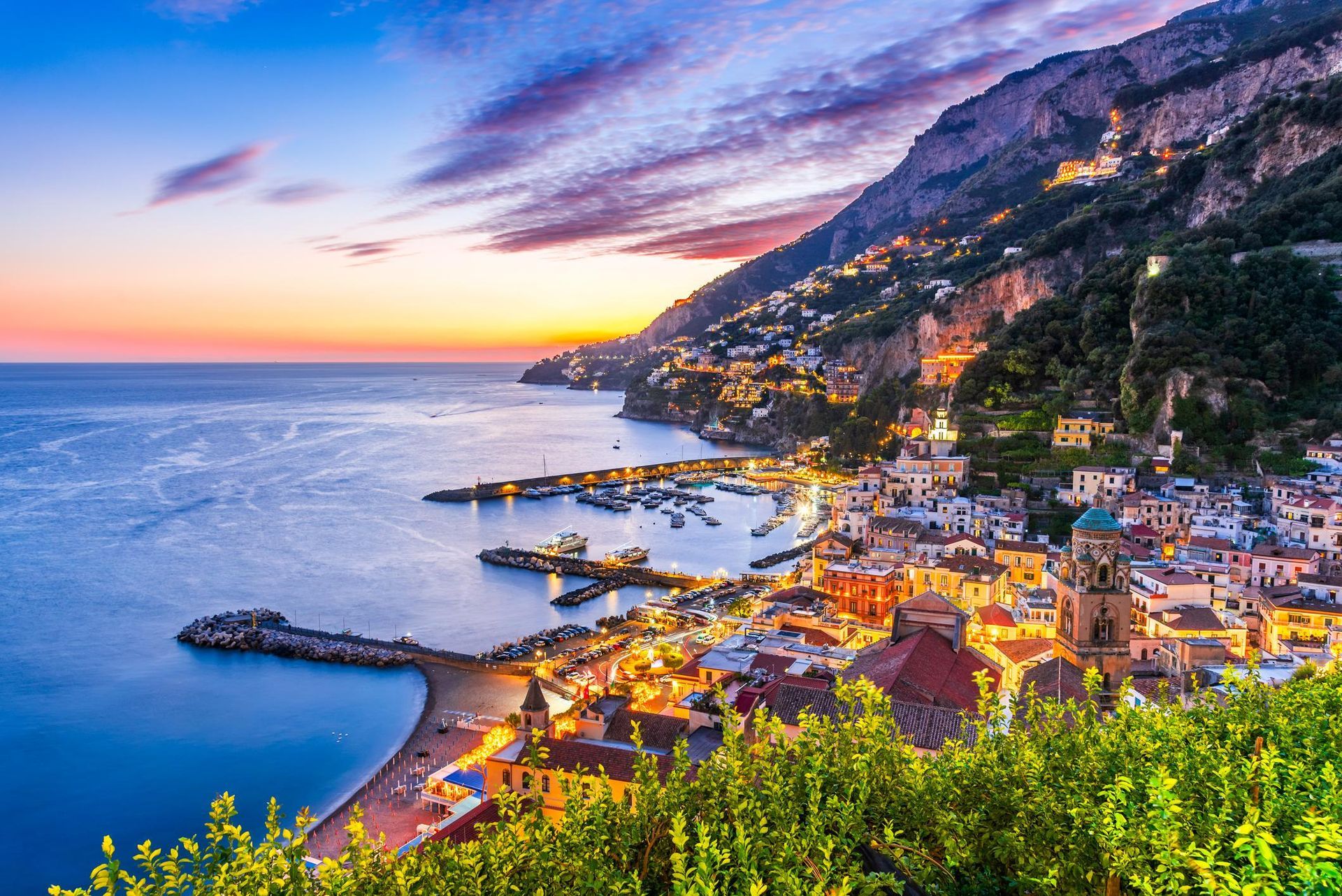 A twilight view of the Amalfi Coast in Italy, with a harbor, town lights, and mountains under a colorful sunset sky.