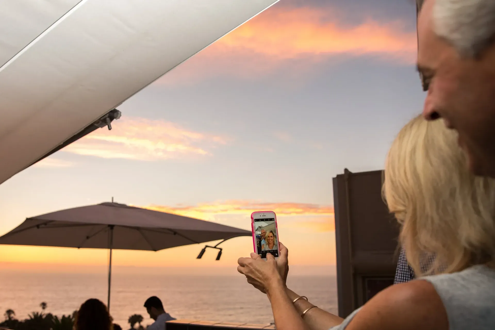 A person holds a phone to capture a sunset over the ocean from a balcony with umbrellas, beside someone looking on.
