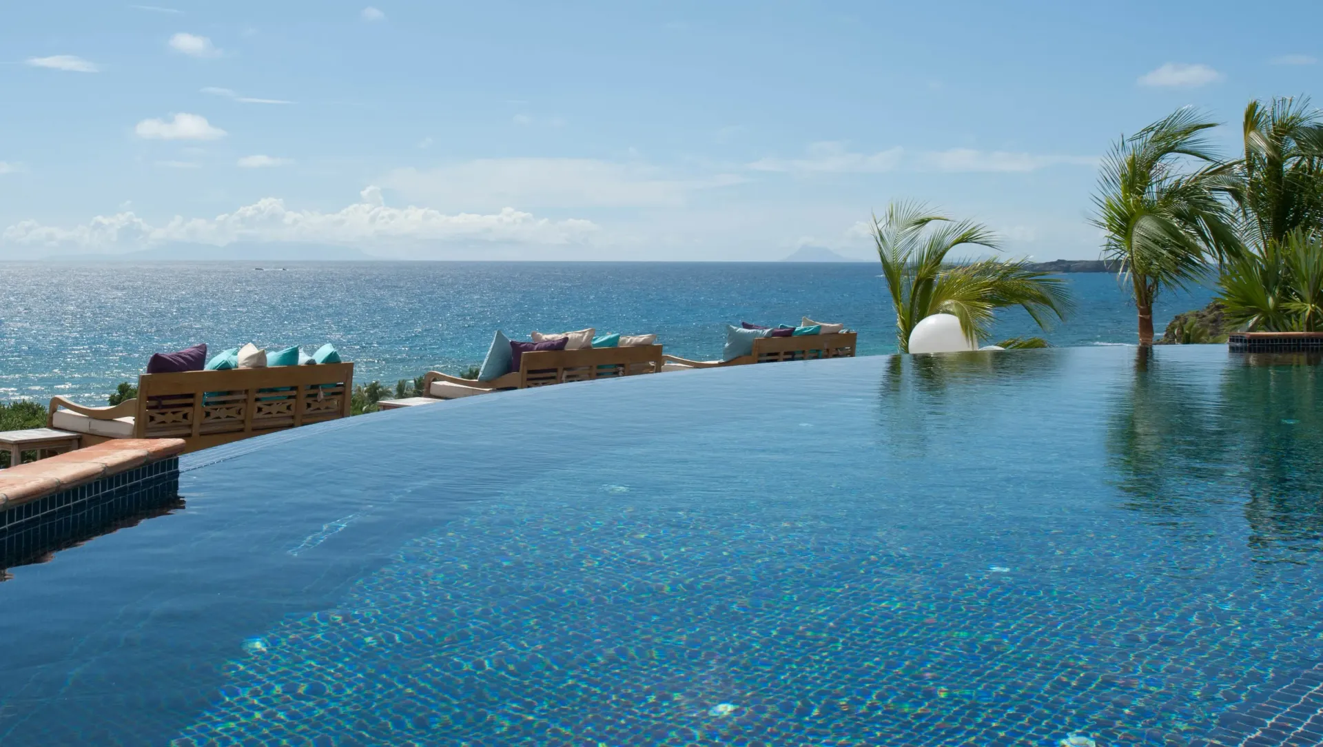 An infinity pool overlooking a sparkling blue ocean under a clear sky, with lounge seating on the edge.