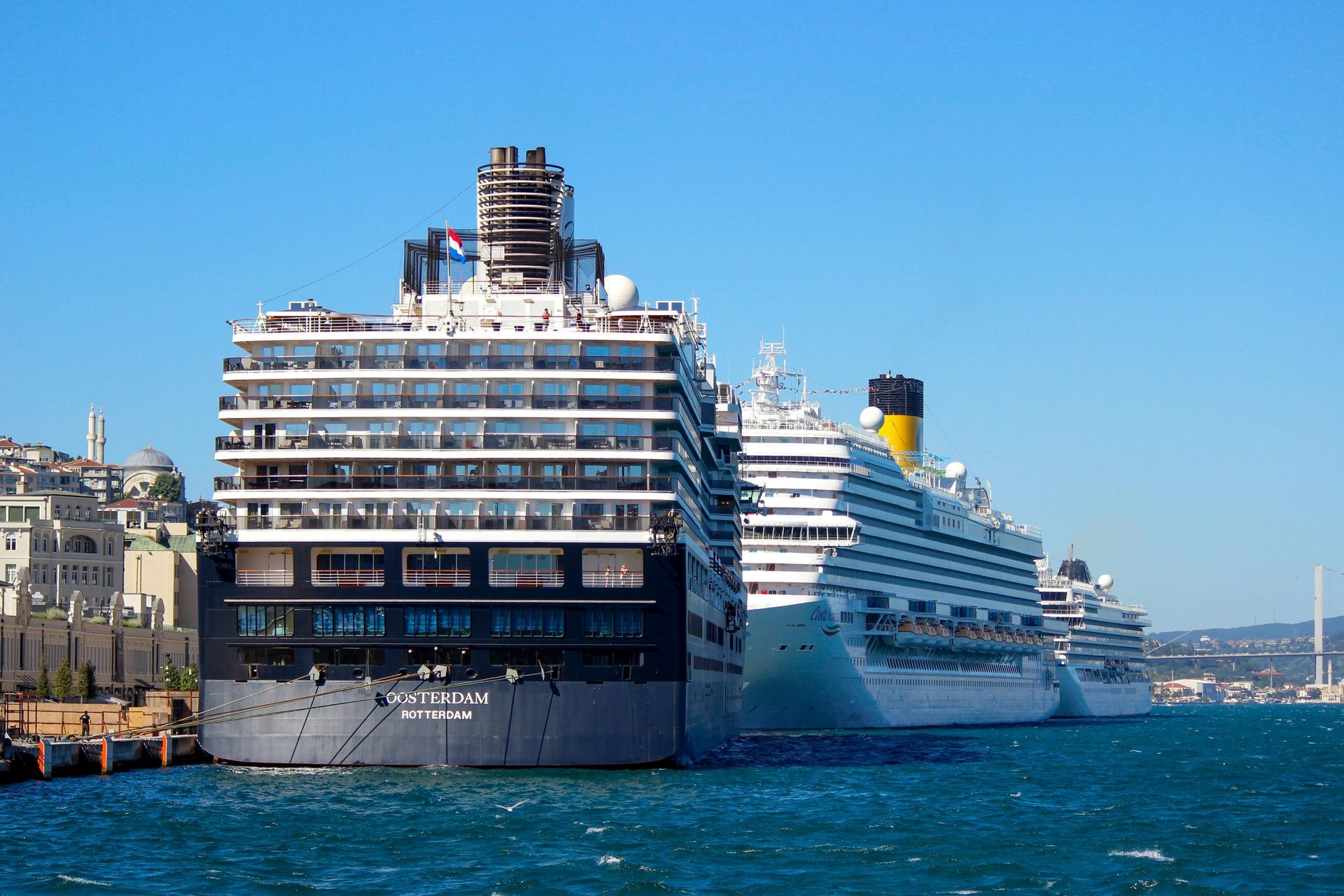 Three large cruise ships are docked in a harbor against a clear blue sky, with a city visible on the left shore.