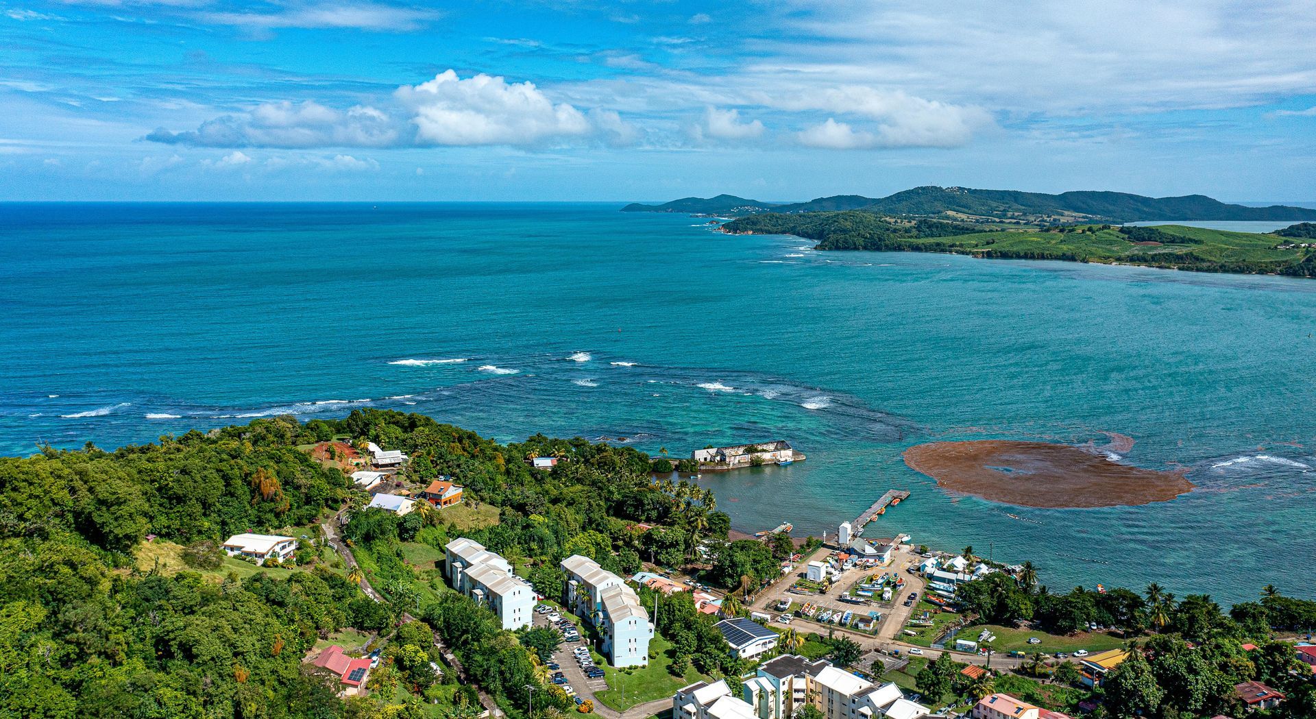An aerial view of a green, tree-covered coastline with white buildings, overlooking a blue ocean with a dark sandy patch.
