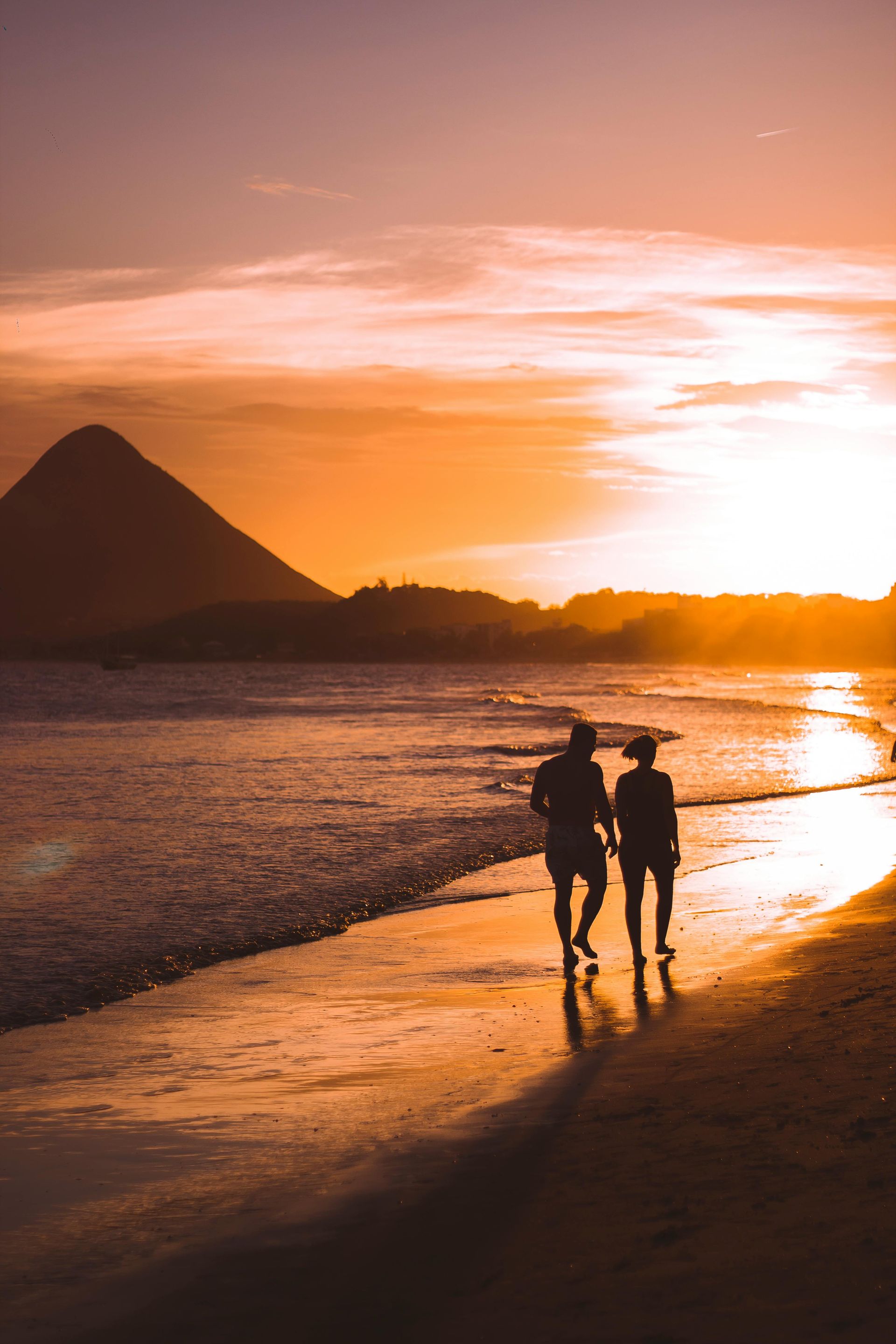 Silhouetted figures walk along a tropical beach at sunset, with a prominent mountain peak in the distance.