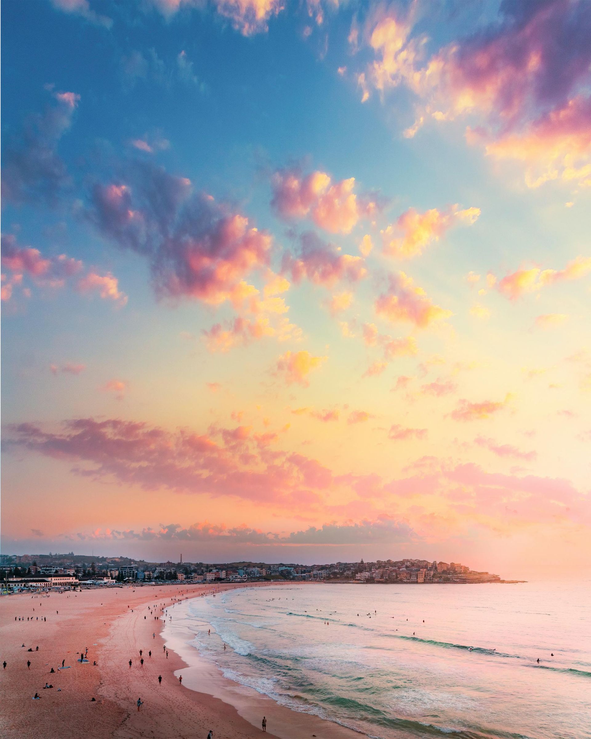 A wide, sandy beach at sunset, with soft pink and orange clouds above a calm ocean and distant coastal buildings.