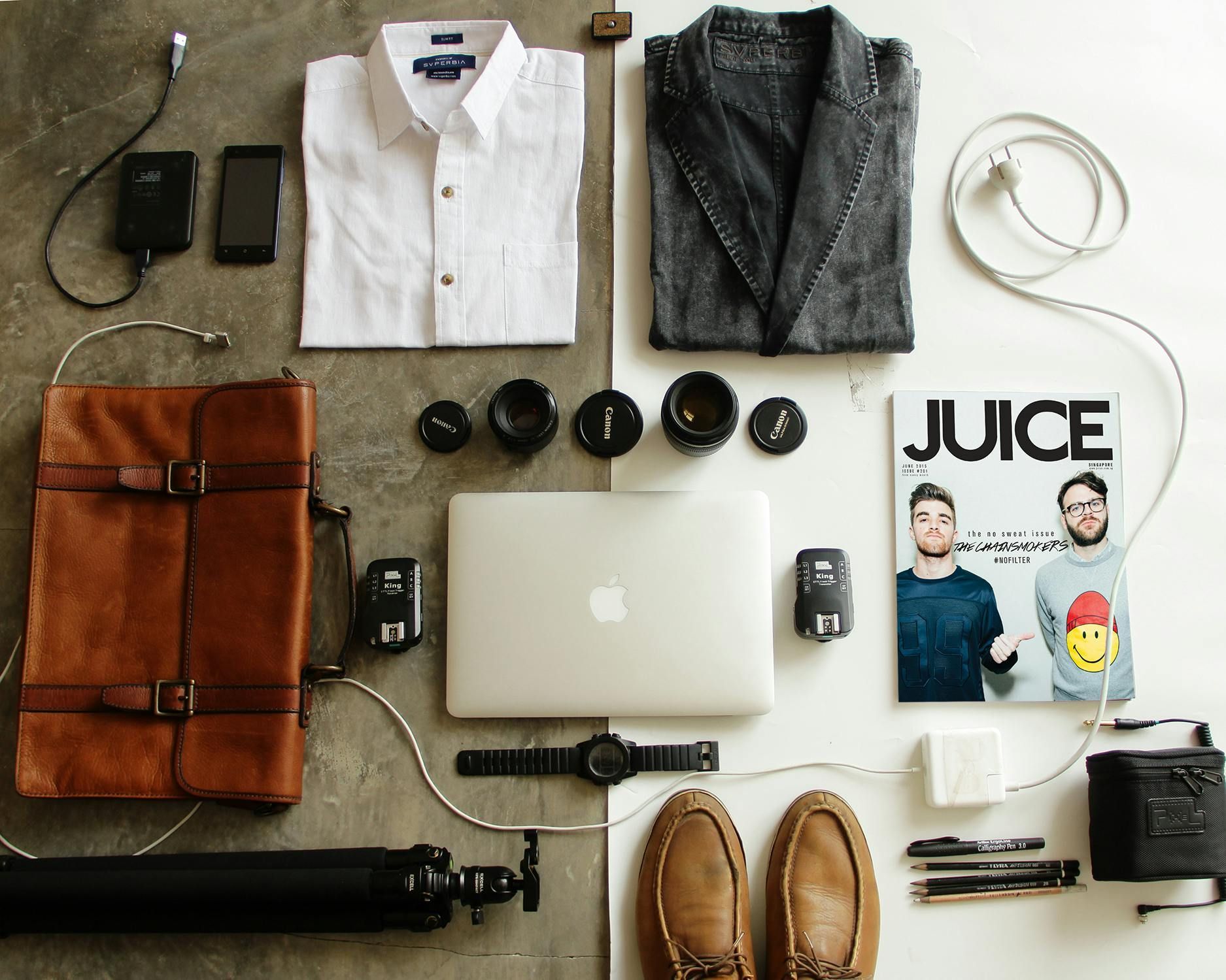 A flat lay of clothing, tech gadgets, camera lenses, a laptop, shoes, and a magazine arranged on a two-toned surface.