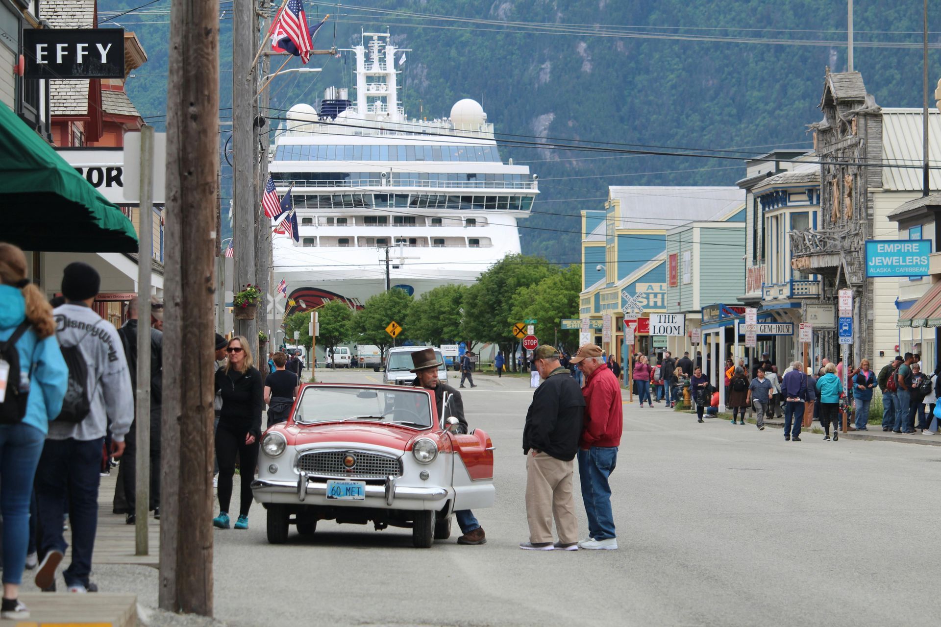 A vintage car parked on a street in front of a large cruise ship docked in a town with buildings and pedestrians.