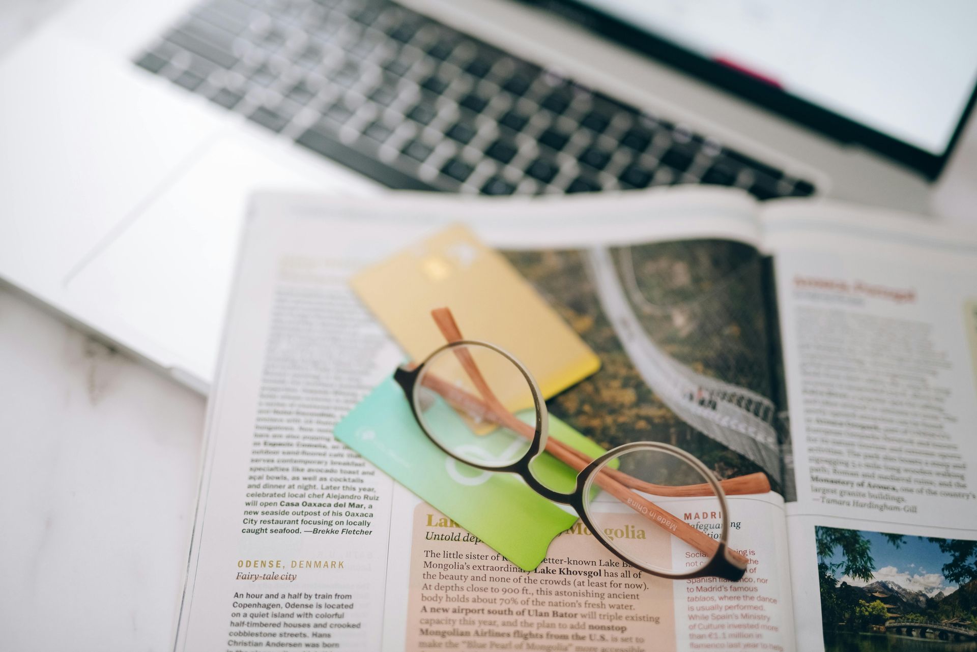 Eyeglasses on an open magazine beside sticky notes and a laptop keyboard