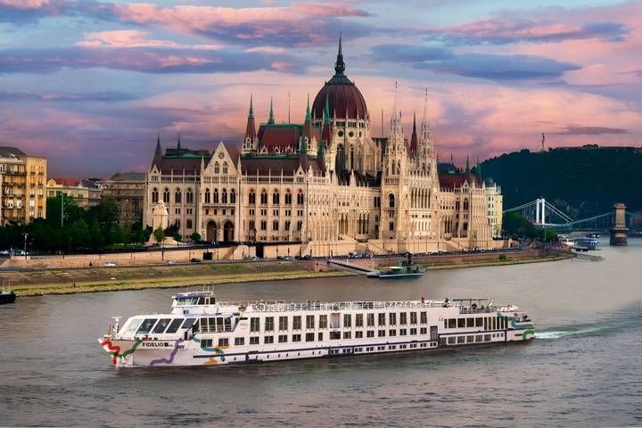 A white river cruise ship travels on the Danube in Budapest, passing the Hungarian Parliament Building at sunset.