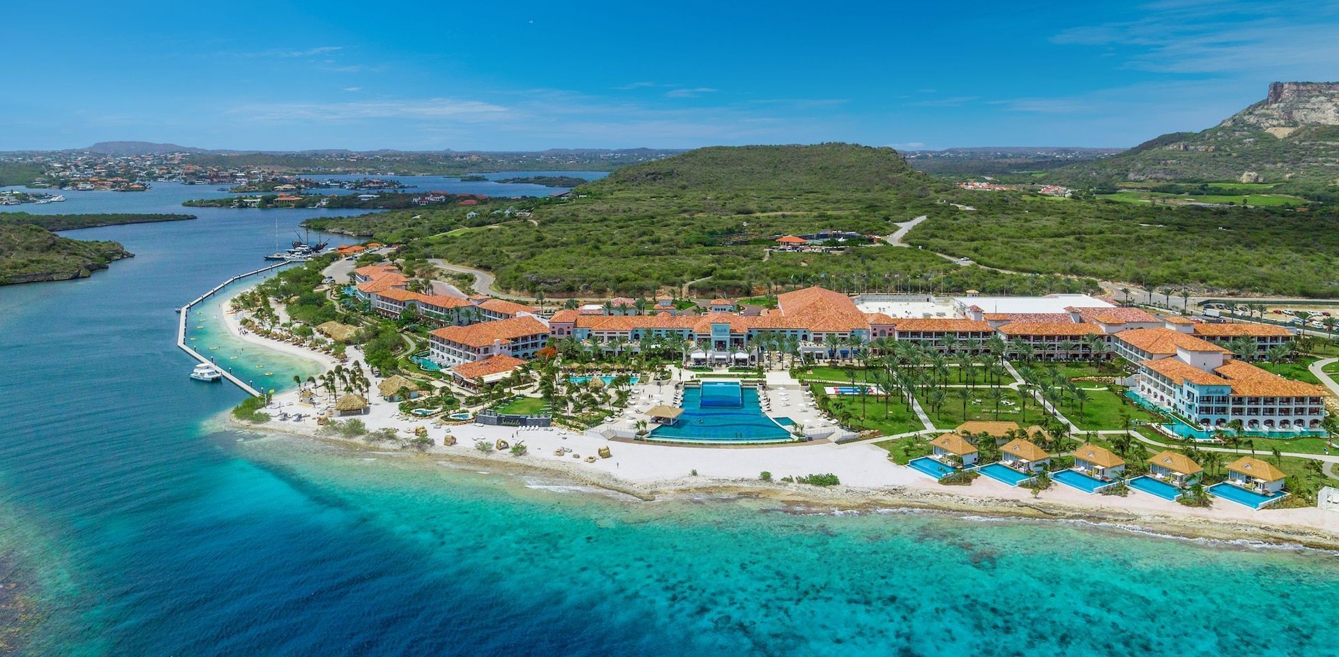 An aerial view of a coastal resort featuring terracotta roofs, swimming pools, white sand beaches, and turquoise water.
