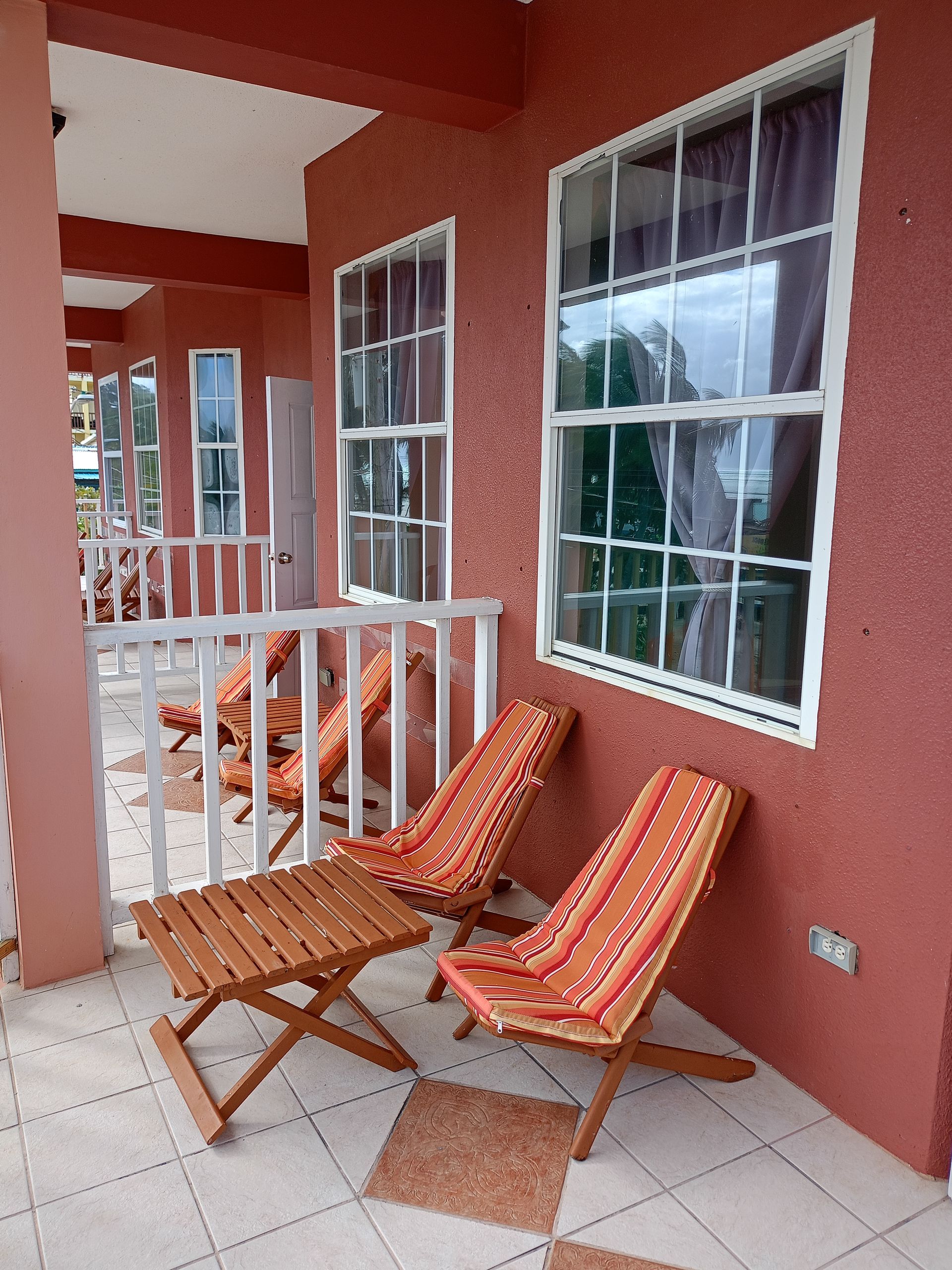 Balcony with two orange-cushioned chairs, a small wooden table, and two windows against a red wall.