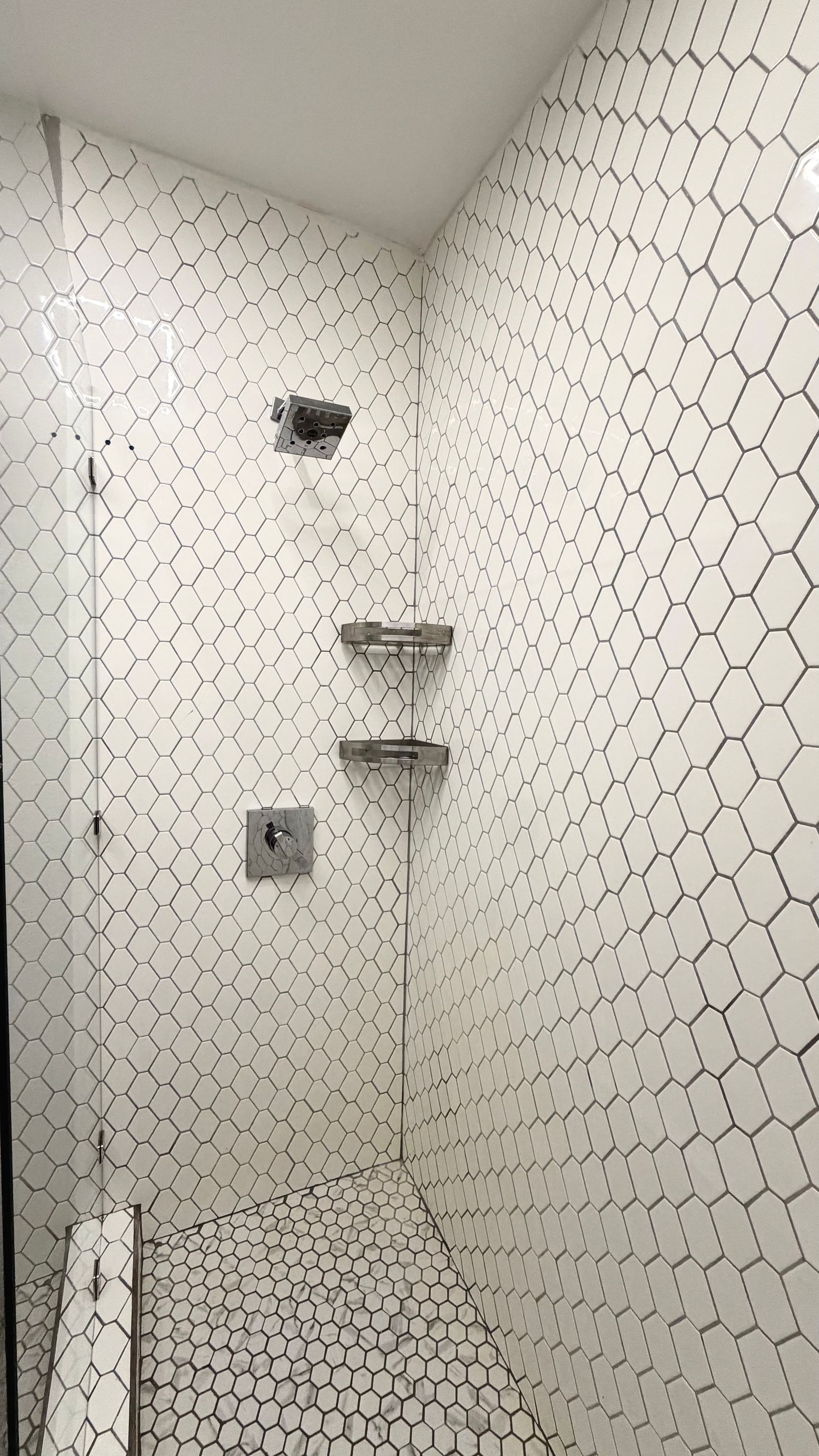 Small tiled shower stall with a corner shelf and wall-mounted shower fixtures.