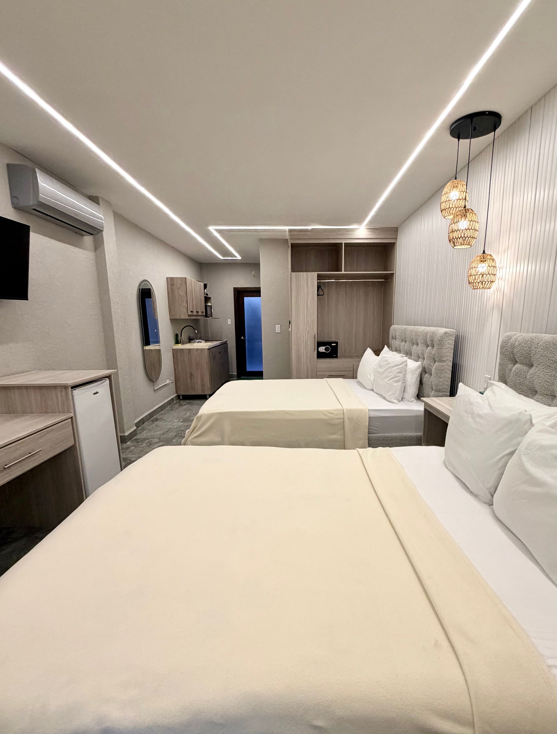 Modern hotel room with two beds, warm pendant lights, wall AC, and a TV on the left.