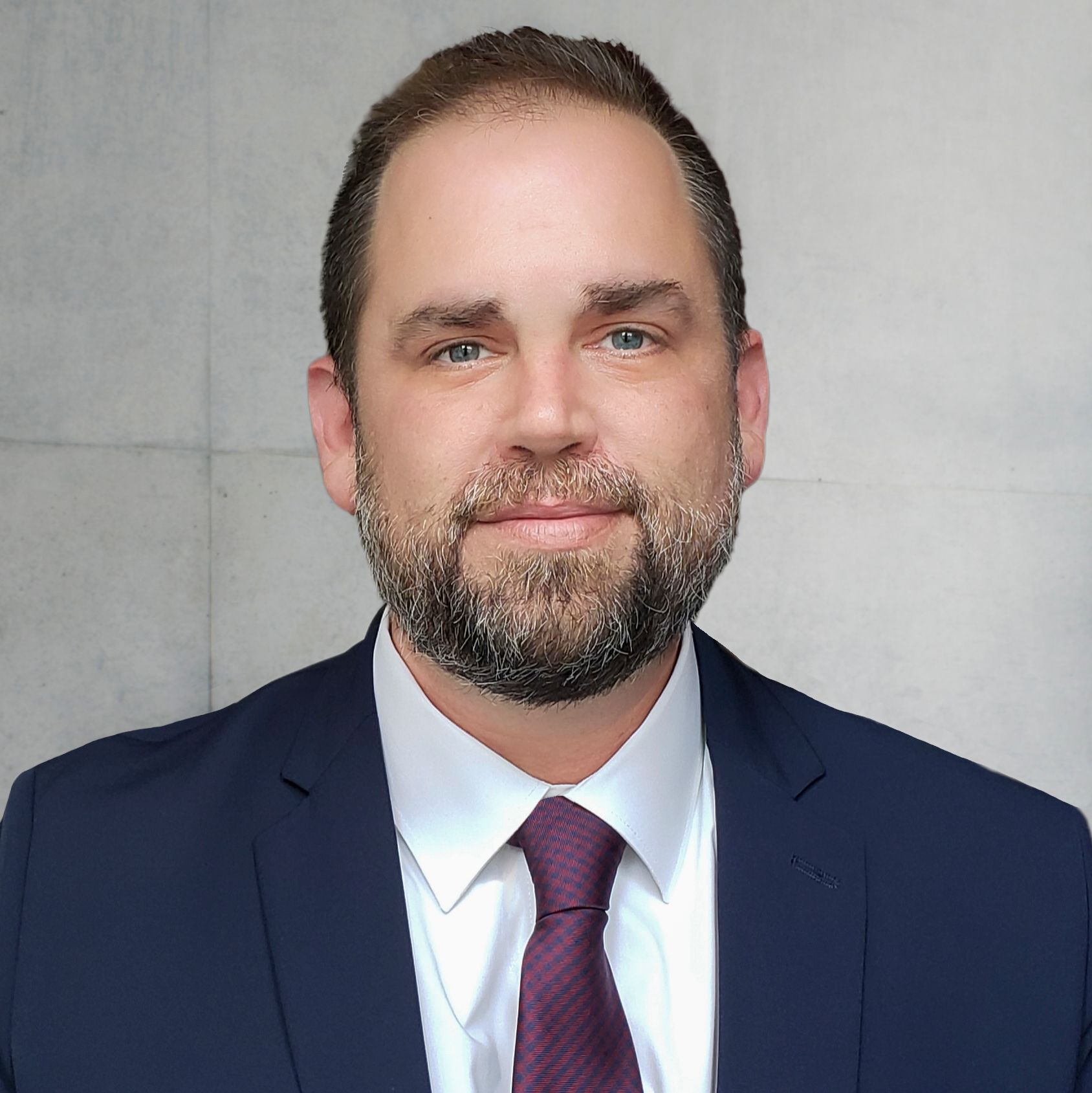 Man in a navy suit, purple tie, beard, and neutral expression, set against a light gray wall.