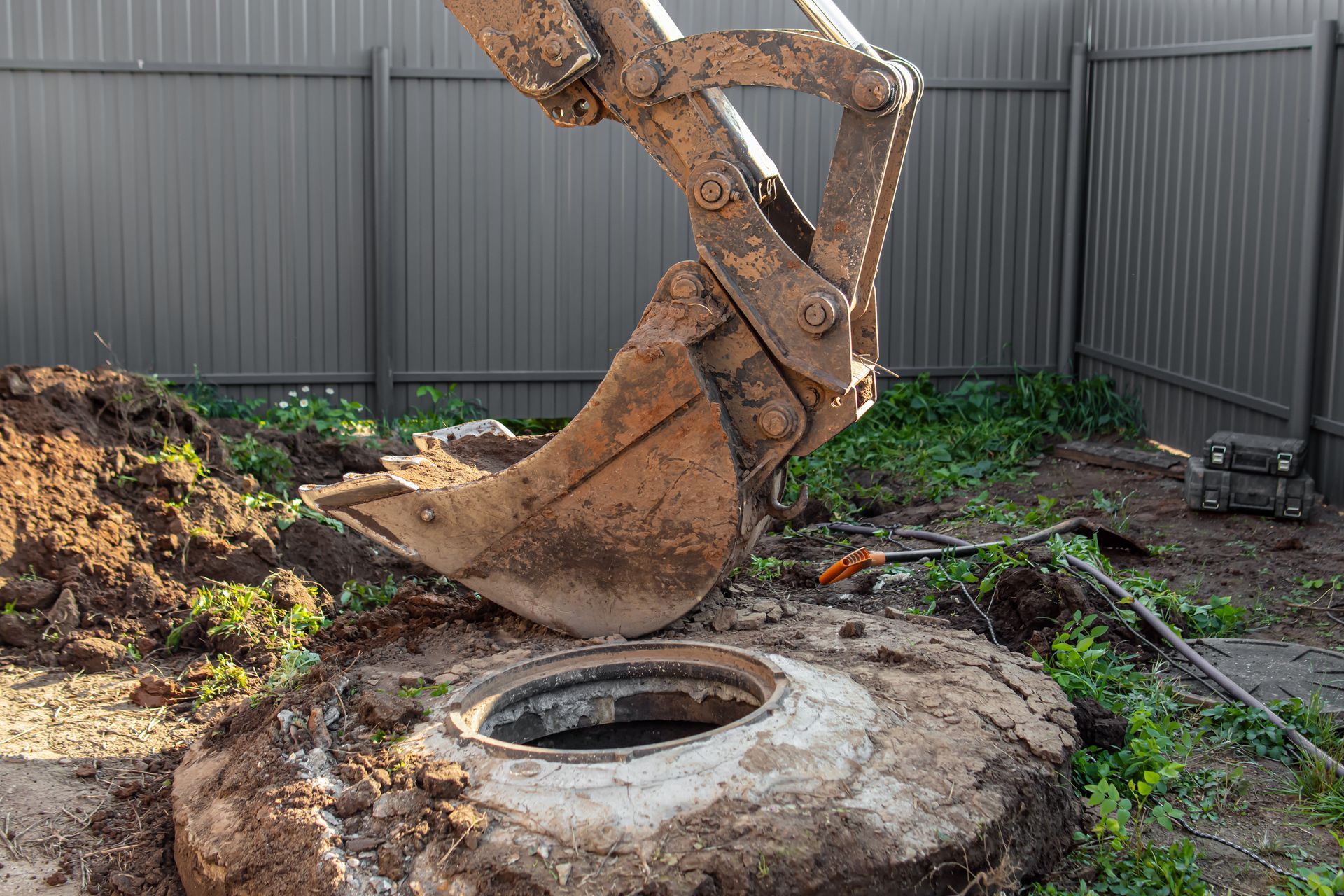 Excavator bucket digging beside a concrete manhole ring in a muddy yard with a metal fence background Excavator bucket digging beside a concrete manhole ring in a muddy yard with a metal fence background