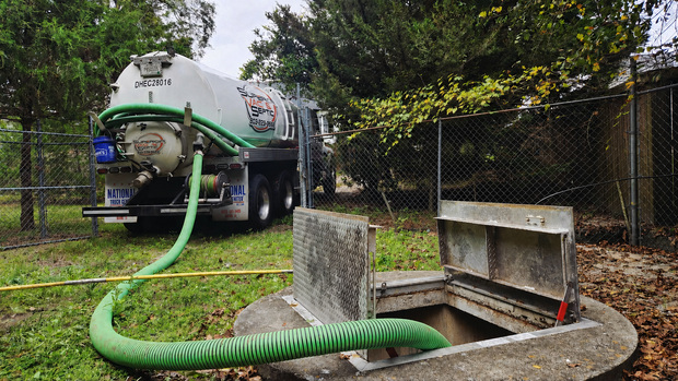 Worker in a neon safety jacket kneels beside a cement truck, inspecting a manhole in a grassy area.