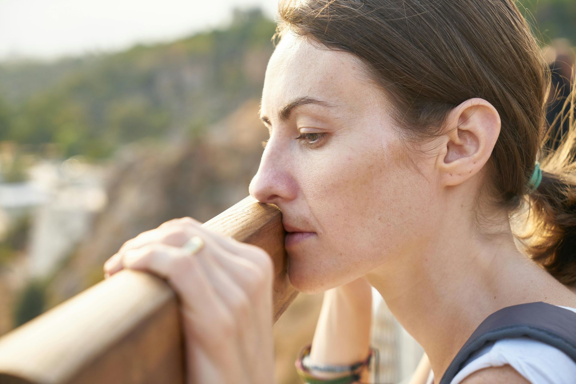 Woman resting chin on wooden railing, looking down with a thoughtful expression. Outdoors, sunny.
