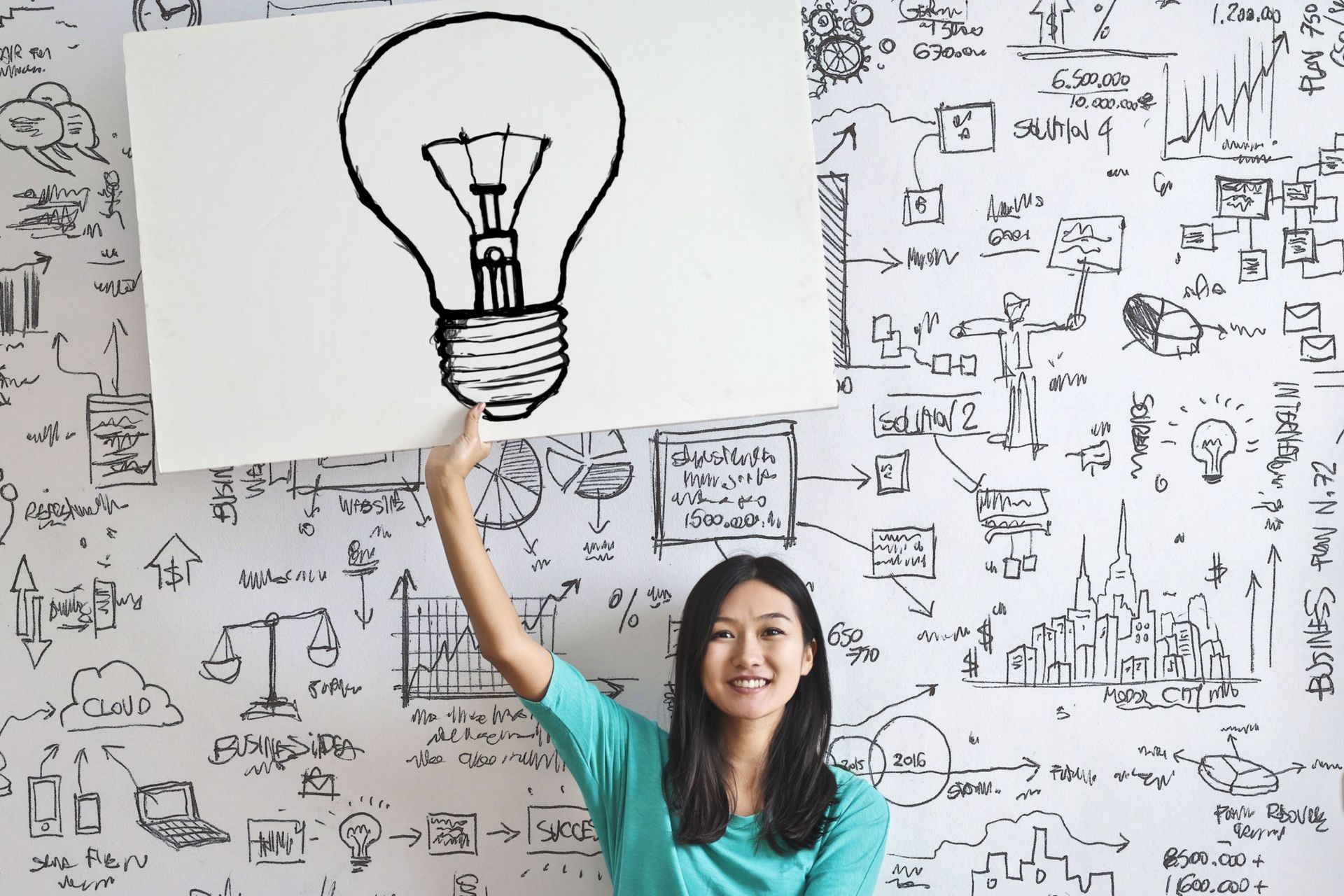 Woman holding a sign with a lightbulb sketch in front of a wall covered in ideas and doodles