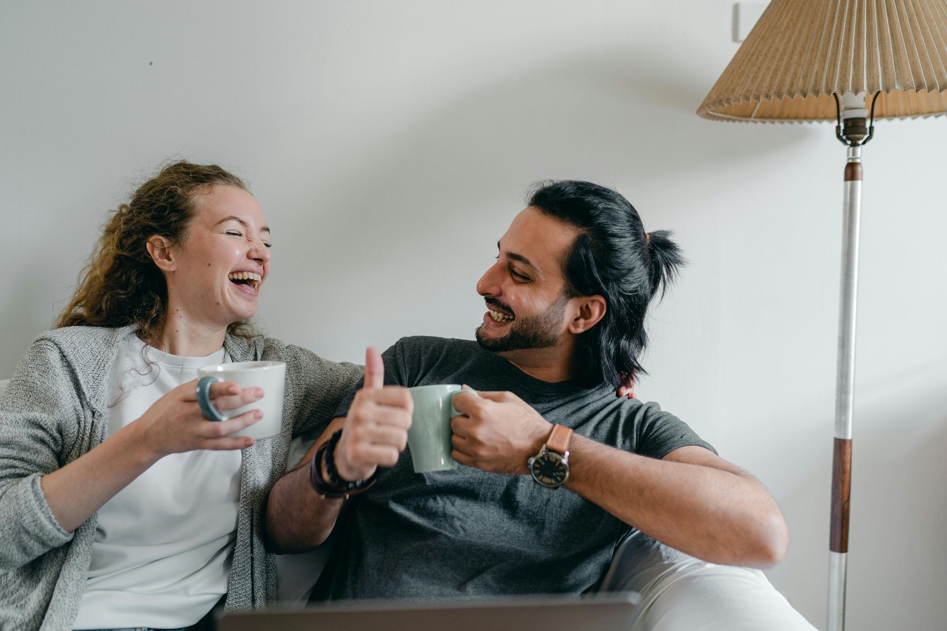 Woman and man laugh, holding mugs. Man gives thumbs up, by a floor lamp.
